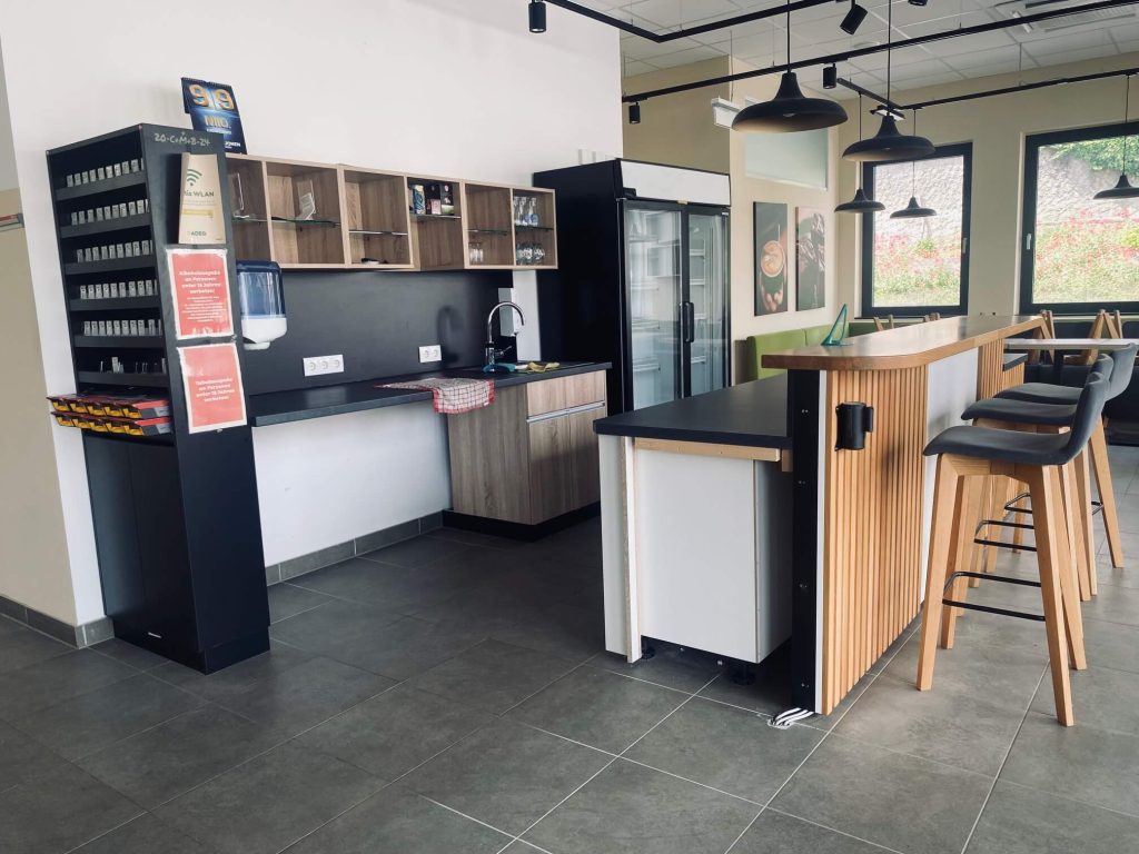 Gesamtansicht eines modernen Kaffeehaus-Thekenbereichs mit Rückwand, Barhockern und Kühleinheit.